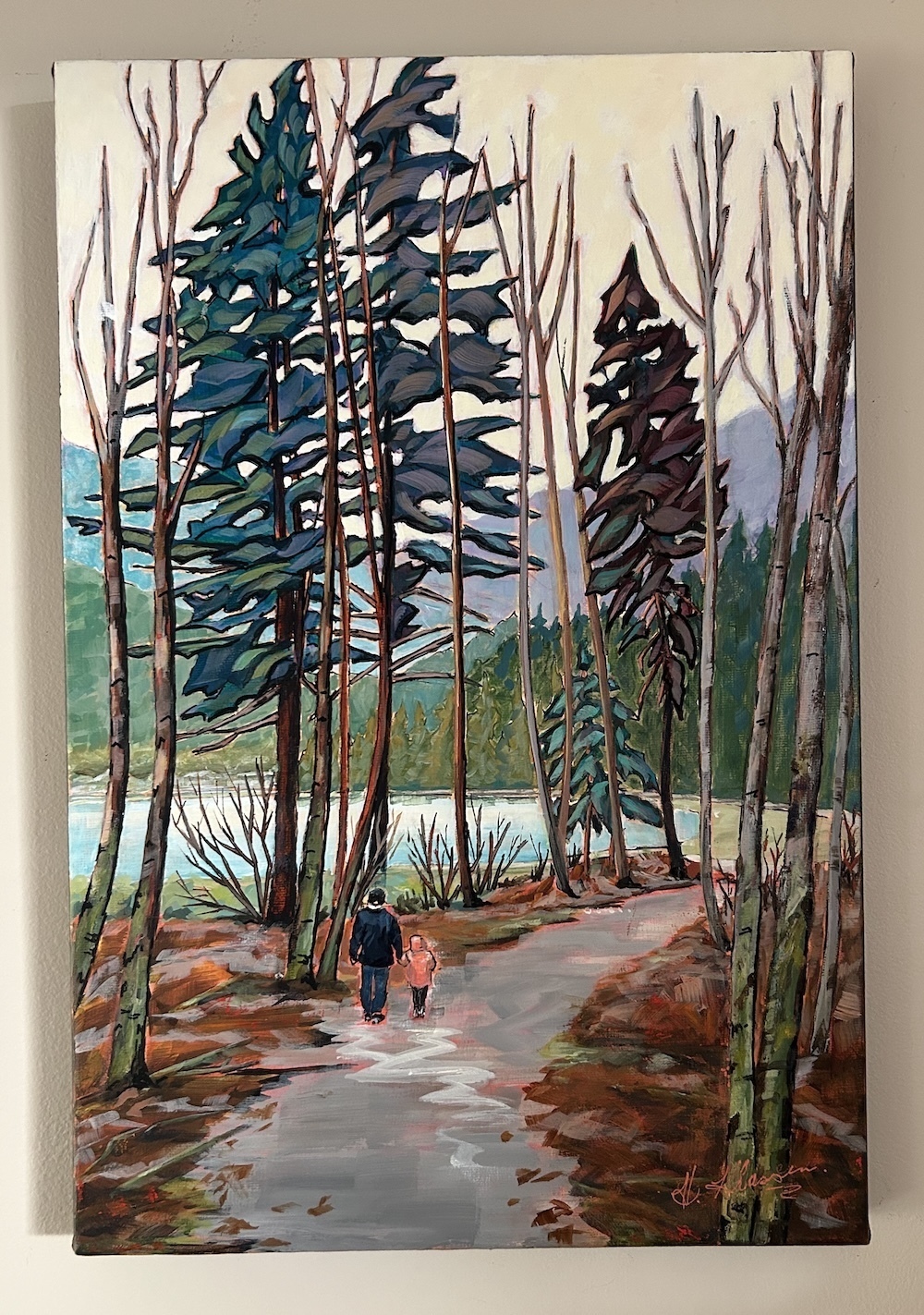 A wooded path beside a lake with distant mountains. An adult and child walk hand in hand on the path